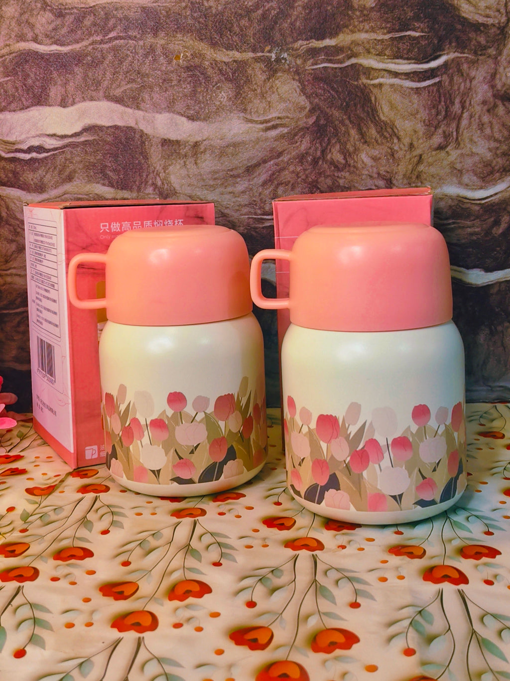 Two floral-patterned thermosfood jars  with pink lids on a floral tablecloth.