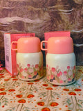 Two floral-patterned thermosfood jars  with pink lids on a floral tablecloth.