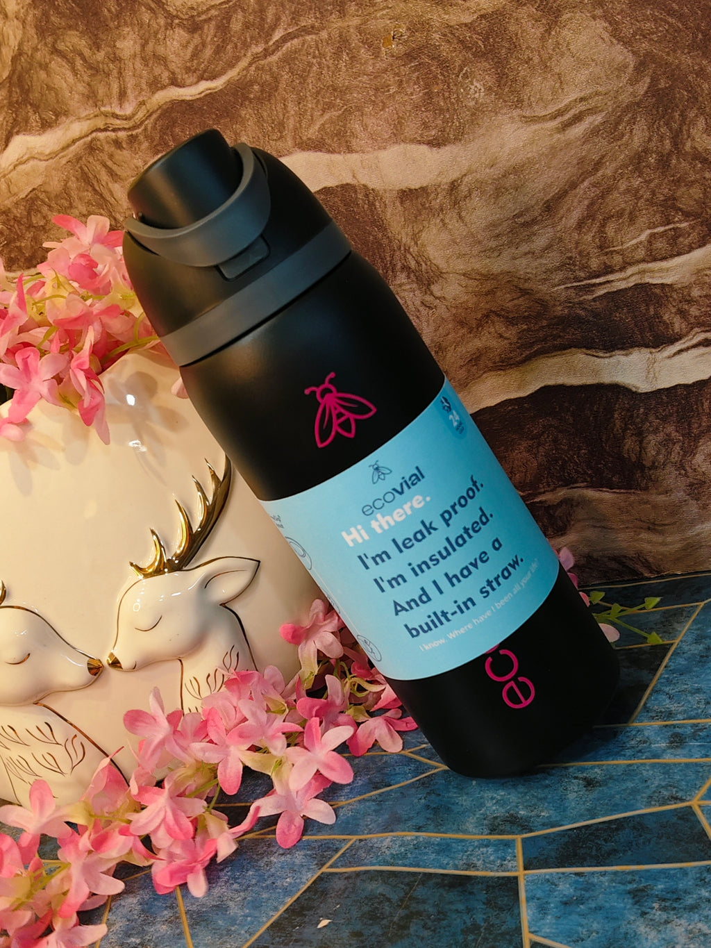 Black insulated bottle with a blue label on a decorative surface with flowers and a deer figurine.
