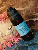 Black insulated bottle with a blue label on a decorative surface with flowers and a deer figurine.