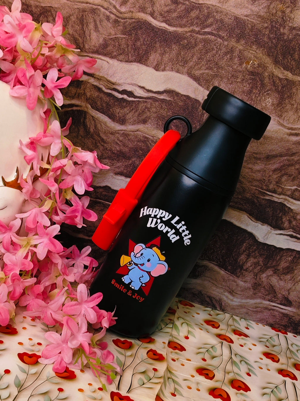 Black water bottle with red cap and 'Happy Little World' branding next to pink flowers on a floral-patterned surface.