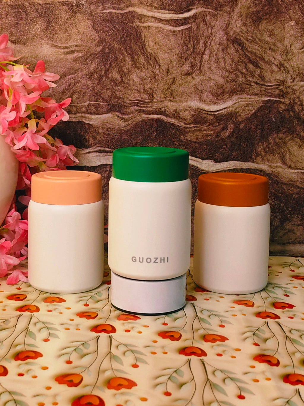 Three white containers with colored lids on a floral-patterned surface with a marble background