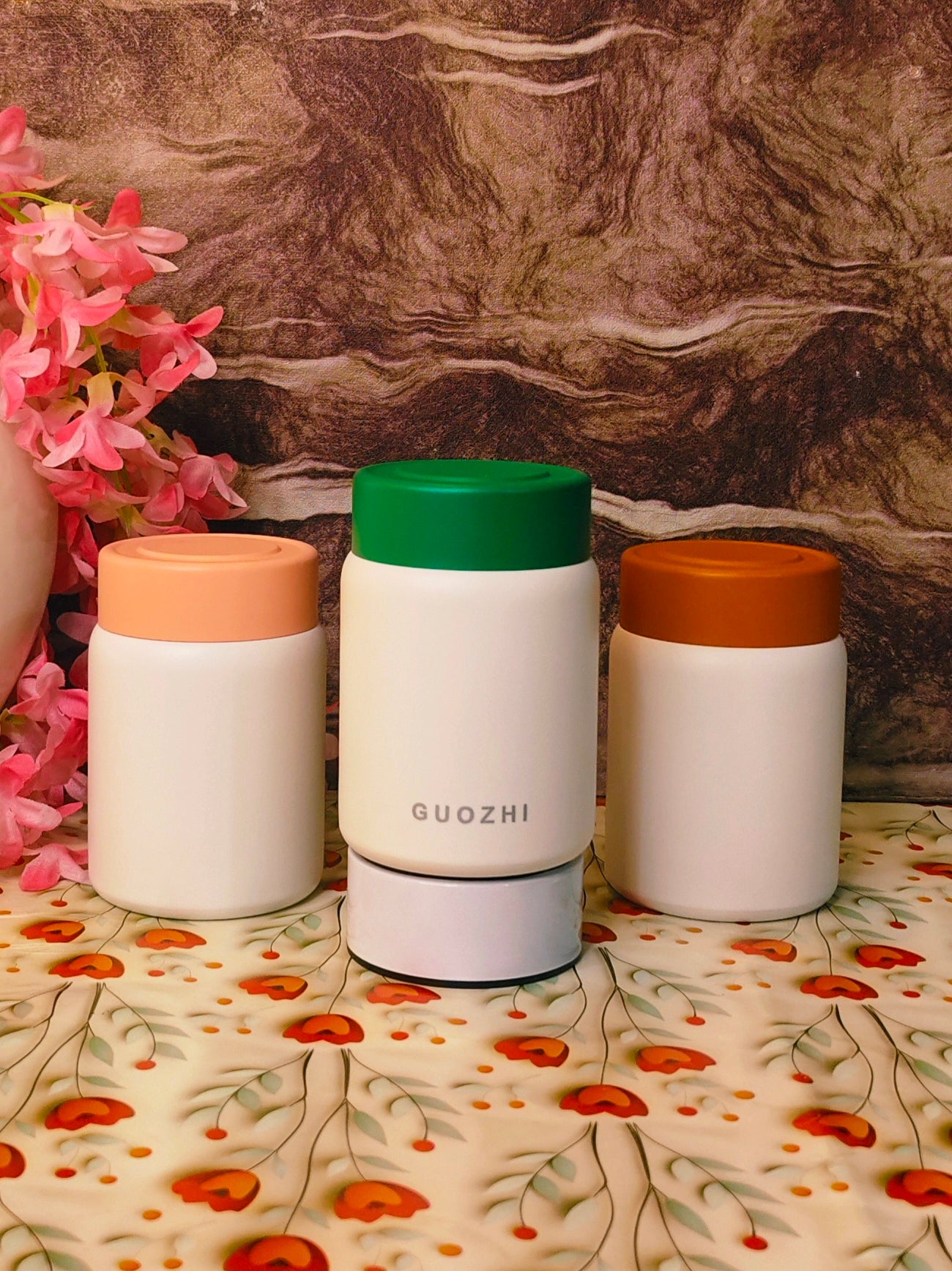 Three white containers with colored lids on a floral-patterned surface with a marble background