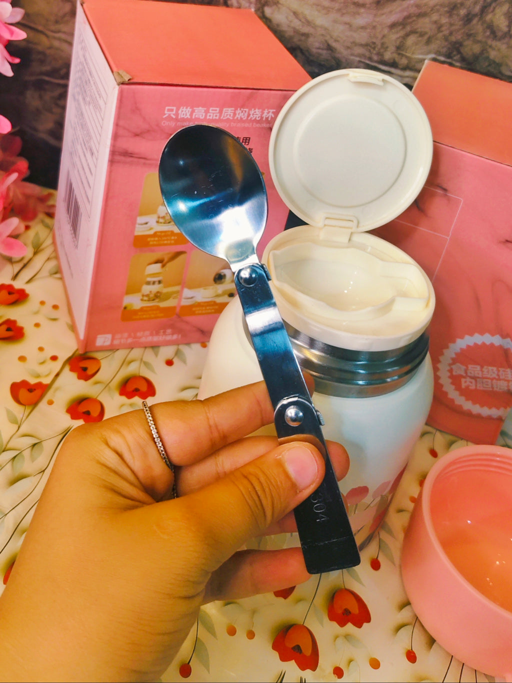 Hand holding a spoon over an open container with a floral and product packaging background